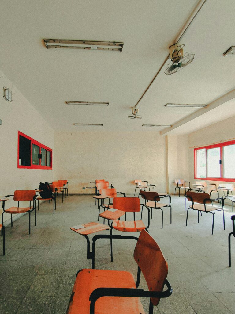 Bright classroom interior featuring empty wooden chairs and desks arranged in rows with red window frames.